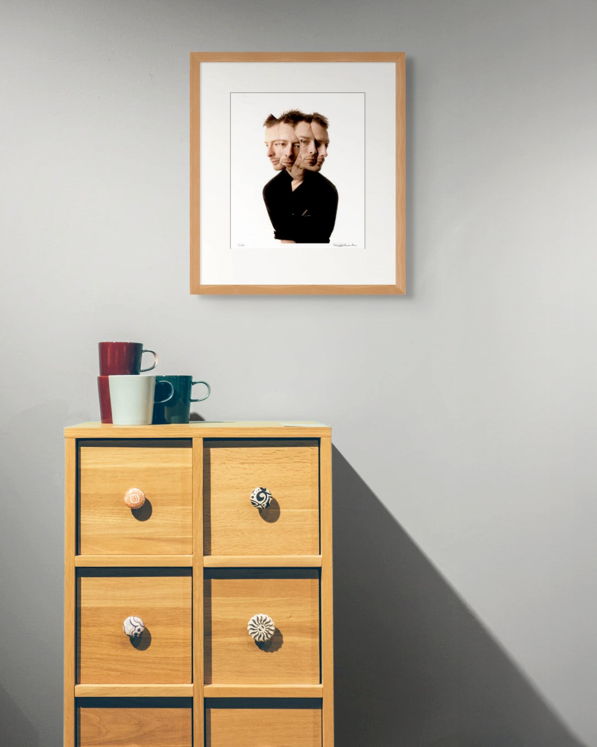Framed Thom Yorke portrait by Robin Sellick above a wooden cabinet with colourful mugs – casual, art-driven home interior styling.